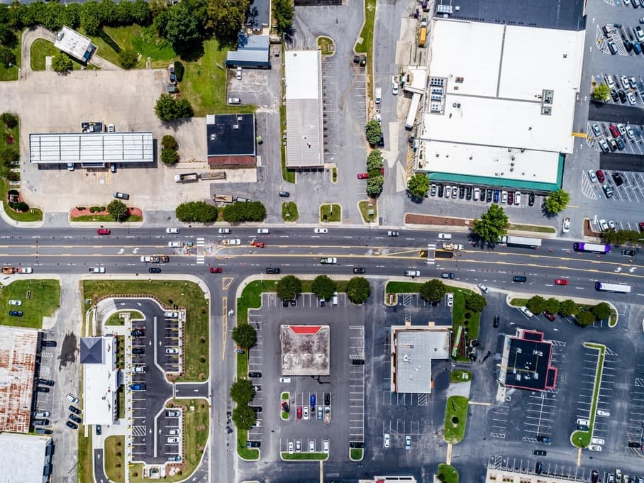 Vertical aerial photograph of a commercial corridor: a large single-span retail roof and its parking field on one side of a divided arterial road, smaller freestanding shop buildings and marked parking bays on the other.