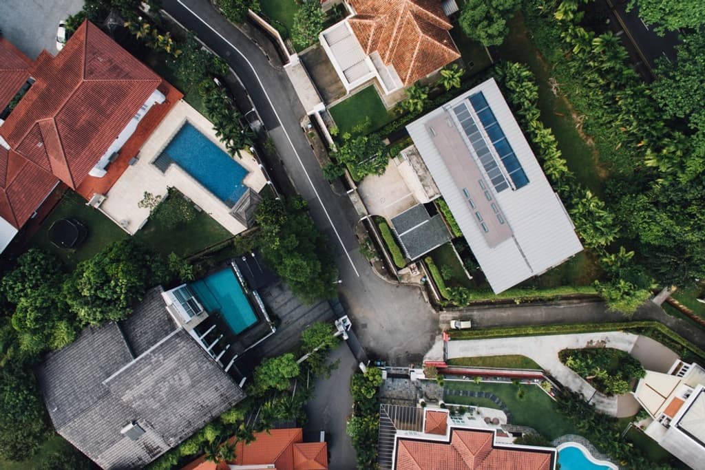 Vertical aerial photograph of a residential block: a long pale roof carrying a rooftop solar array, a large dark-tiled roof with a pool beside it, and a red-tiled roof partly under tree canopy, each on its own lot.